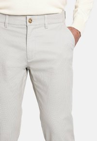 Hombre con pantalones texturizados en gris claro y la mano en el bolsillo, combinado con una camiseta de manga larga color crema.