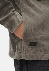 Persoon die een grijze ribfluweel jasje draagt met de hand in de zak, met een klein zwart en geel "camel active" label dicht bij de zoom.