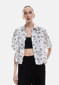 Camicia bianca a maniche corte con bottoni, decorata con motivi di draghi neri e fiamme, abbinata a un crop top nero e pantaloni neri a vita alta.