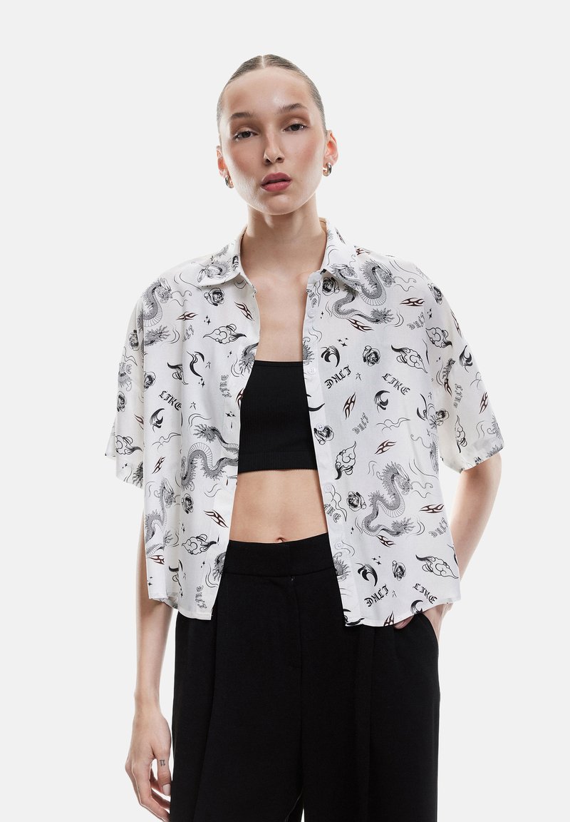 Camicia bianca a maniche corte con bottoni, decorata con motivi di draghi neri e fiamme, abbinata a un crop top nero e pantaloni neri a vita alta.