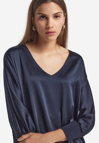 Marineblaue Satinbluse mit V-Ausschnitt, langen Puffärmeln und elastischen Bündchen. Glatte Textur mit einem dezenten Glanz, entworfen für eine lockere Passform.