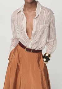 Vrouw in een beige gestreepte shirt, ingestopt in een roestkleurige rok met een bruine riem en chunky gouden en zwarte armbanden.