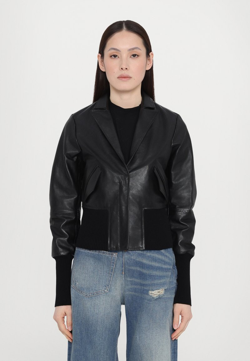 Black leather jacket with a fitted waist, wide lapels, and ribbed cuffs. Worn over a black top and paired with light blue distressed jeans.