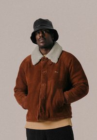 Chaqueta de ante marrón con cuello de borrego crema, cierre de botones y bolsillos laterales. Sombrero bucket negro complementa el conjunto.