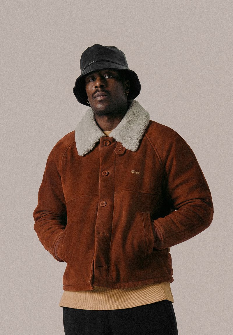 Chaqueta de ante marrón con cuello de borrego crema, cierre de botones y bolsillos laterales. Sombrero bucket negro complementa el conjunto.