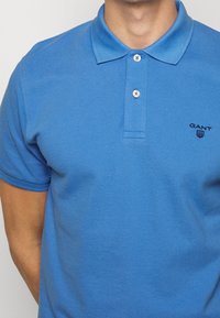 Modrá bavlněná polo košile s texturovanou tkaninou, tříknoflíkovou légou a malým černým logem na levém prsu. Krátké rukávy.