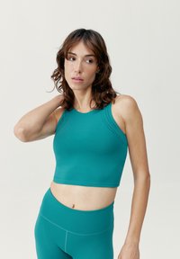 Tyrkysový sportovní crop top s vysokým výstřihem, zdobený perforovanými detaily na ramenou a hladkým, elastickým materiálem. V kombinaci s odpovídajícími legínami.