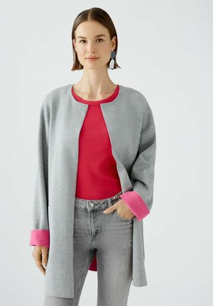 Light gray cardigan with pink accents, worn over a red ribbed top. The model is in gray jeans, showcasing a casual, layered look.