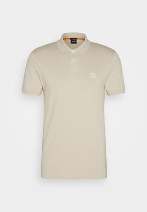 Beige polo shirt made of soft fabric, featuring a collar, three buttons, and a logo patch on the left chest. Short sleeves and a clean design.