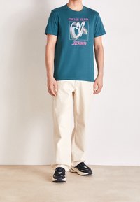 Turkusowa bawełniana koszulka z grafiką "Calvin Klein Jeans" w różowym i białym kolorze, zestawiona z beżowymi spodniami i czarnymi sneakersami, stojąca na neutralnej podłodze.