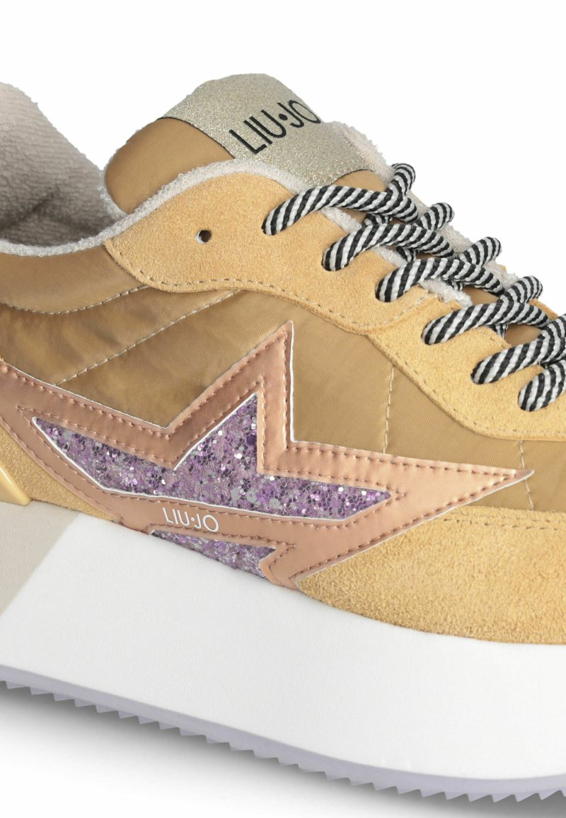 Zapatilla con una parte superior de ante y nailon beige, que presenta un detalle de estrella morada brillante, cordones de rayas negras y blancas, y una suela gruesa blanca.