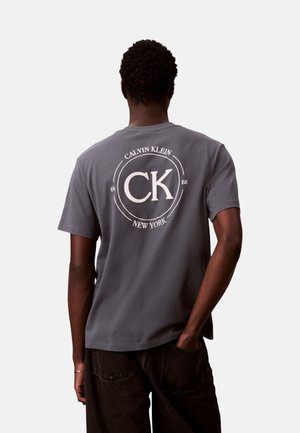 Uomo che indossa una t-shirt grigia Calvin Klein con logo bianco rotondo e testo "CK Calvin Klein New York 1968" sul retro, pantaloni neri, mano in tasca.