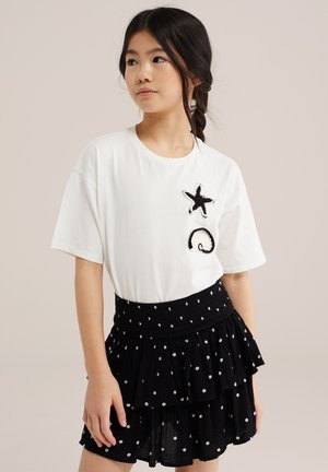 Mladé dievča s tmavými vrkočmi, oblečené v bielej blúzke so záplatami hviezdíc a mušlí a v čiernej sukni s viacvrstvovým volánom s bodkami.