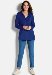 Marineblaue Bluse mit langen Ärmeln, Kragen und vorderen Falten, kombiniert mit hellblauen, hochgekrempelten Jeans und weißen Sneakers.