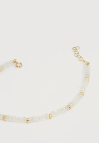 Λεπτό βραχιόλι με μικρές ημιδιαφανείς λευκές χάντρες και χρυσά διαχωριστικά, με χρυσό κούμπωμα και ρυθμιζόμενη αλυσίδα σε λευκό φόντο.