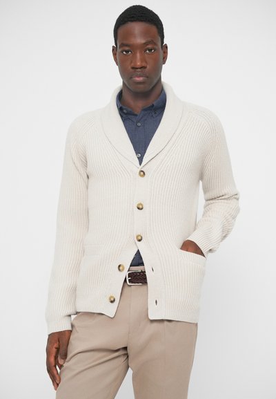 Krém színű, ribb szabású kardigán sálgallérral, elülső gombokkal és oldalzsebekkel, sötétkék inggel és bézs nadrággal párosítva.