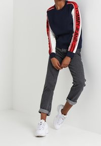Sweat-shirt bleu marine avec manches rayées rouge et blanc, arborant le logo "TOMMY JEANS" ; associé à un jean gris retroussé et des baskets blanches.