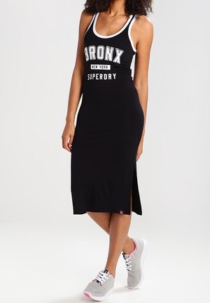 Frau trägt ein schwarzes ärmelloses Midikleid mit dem Schriftzug "BRONX New York SUPERDRY", kombiniert mit grauen und pinken Sneakers, steht mit einem Bein über dem anderen.
