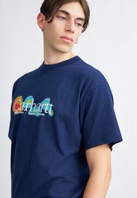 T-shirt in cotone blu navy con stampa grafica multicolore che riporta il nome del brand "Carhartt" e un testo sottostante. Design a maniche corte e collo tondo.