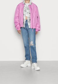 Person som bär en rosa bombarjacka, skjorta med svartvitt serietryck, slitna blå jeans och klumpiga vita plattformssneakers.