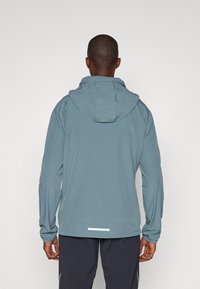 Veste de sport pour homme en bleu, fabriquée en tissu léger, dotée d'une capuche, de poignets élastiques et d'un accent réfléchissant près de l'ourlet.