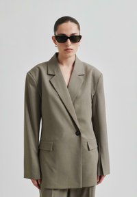 Överdimensionerad kakhifärgad blazer med en svart knapp, skarpa slag, två framfickor och strukturerade axlar, tillsammans med mörka solglasögon.