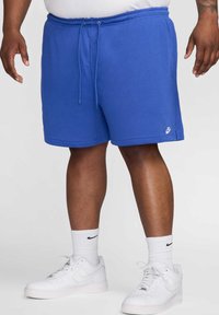 Shorts di cotone blu con vita elasticizzata e cordino. Presentano un piccolo logo Nike bianco sulla gamba sinistra. Indossati con sneakers bianche.