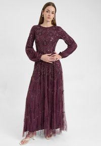 Lungo vestito da sera viola con maniche lunghe, arricchito di paillettes e ricami floreali, sovrapposto a un tessuto trasparente.