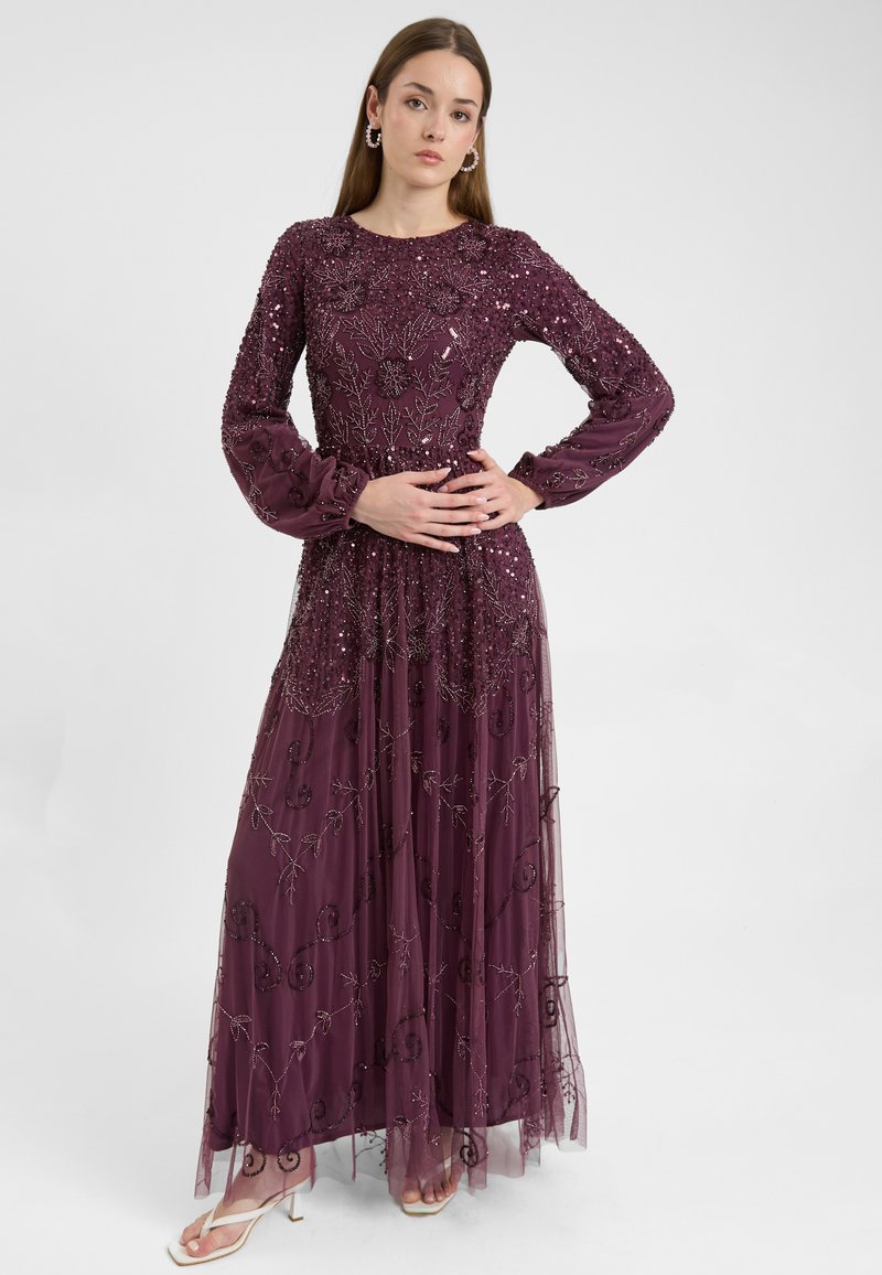 Lungo vestito da sera viola con maniche lunghe, arricchito di paillettes e ricami floreali, sovrapposto a un tessuto trasparente.