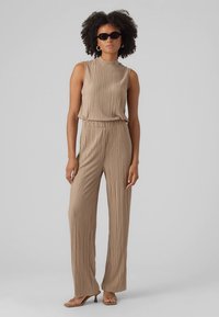 Bež plisiran kombinezon z visokim ovratnikom, sproščenim krojem, širokimi hlačnicami in oprijetim pasom, kombiniran z sončnimi očali in minimalistično obutvijo.