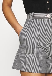 Pantaloni corti a righe bianche e nere con vita alta, caratterizzati da un orlo arrotolato e tasche laterali, chiusi con un bottone tondo.