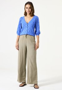 Cardigan bleu brodé avec un col en V et des manches bouffantes, associé à un pantalon en lin beige à jambes larges et des sandales plates noires.