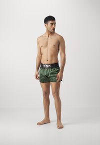 Calções de cetim verdes com faixa elástica preta apresentando o logótipo "VENUM". O modelo está descalço, sem camisa, com tatuagens visíveis no braço.