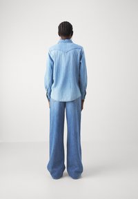 Chemise en denim bleu clair avec un détail de découpe et des manches longues, associée à un pantalon en denim bleu clair à jambes larges. Texture lisse, design décontracté.
