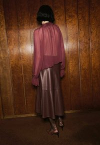 Bordeaux doorzichtige blouse met lange, vloeiende mouwen, gecombineerd met een bruine leren midi-rok. Stiletto-hakken zichtbaar, met patroonsleggingen.