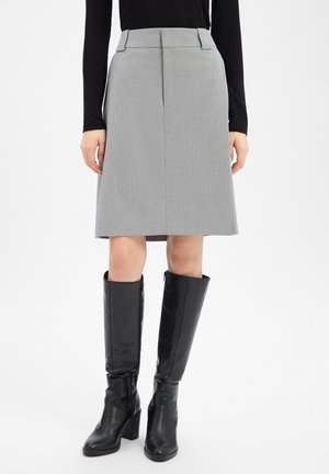 Vrouw draagt een grijze rok tot op de knie en zwarte kniehoge laarzen met hakken, met een zwart topje met lange mouwen, staand tegen een witte achtergrond.