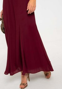 Robe bordeaux longue en tissu léger, avec un design fluide en A. Associée à des escarpins métalliques à lanières et une pochette texturée.