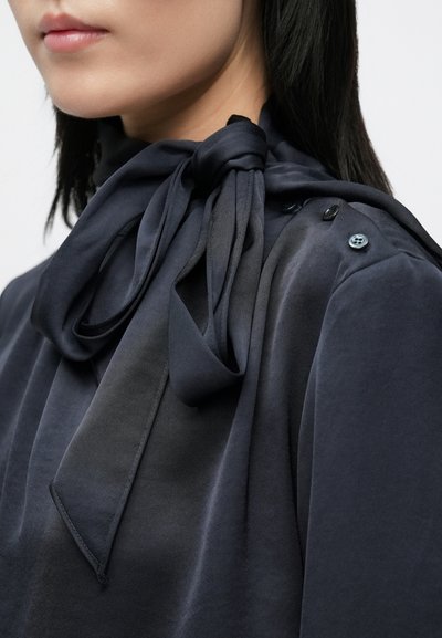 Blusa blu navy scuro con sciarpa annodata al collo, tessuto morbido e bottoni sulla spalla. Tessuto liscio e dettagli di design eleganti.