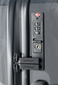 Candado de combinación con cerradura TSA en maleta negra, dos tiradores de cremallera con la marca Off-White asegurados a través del candado.