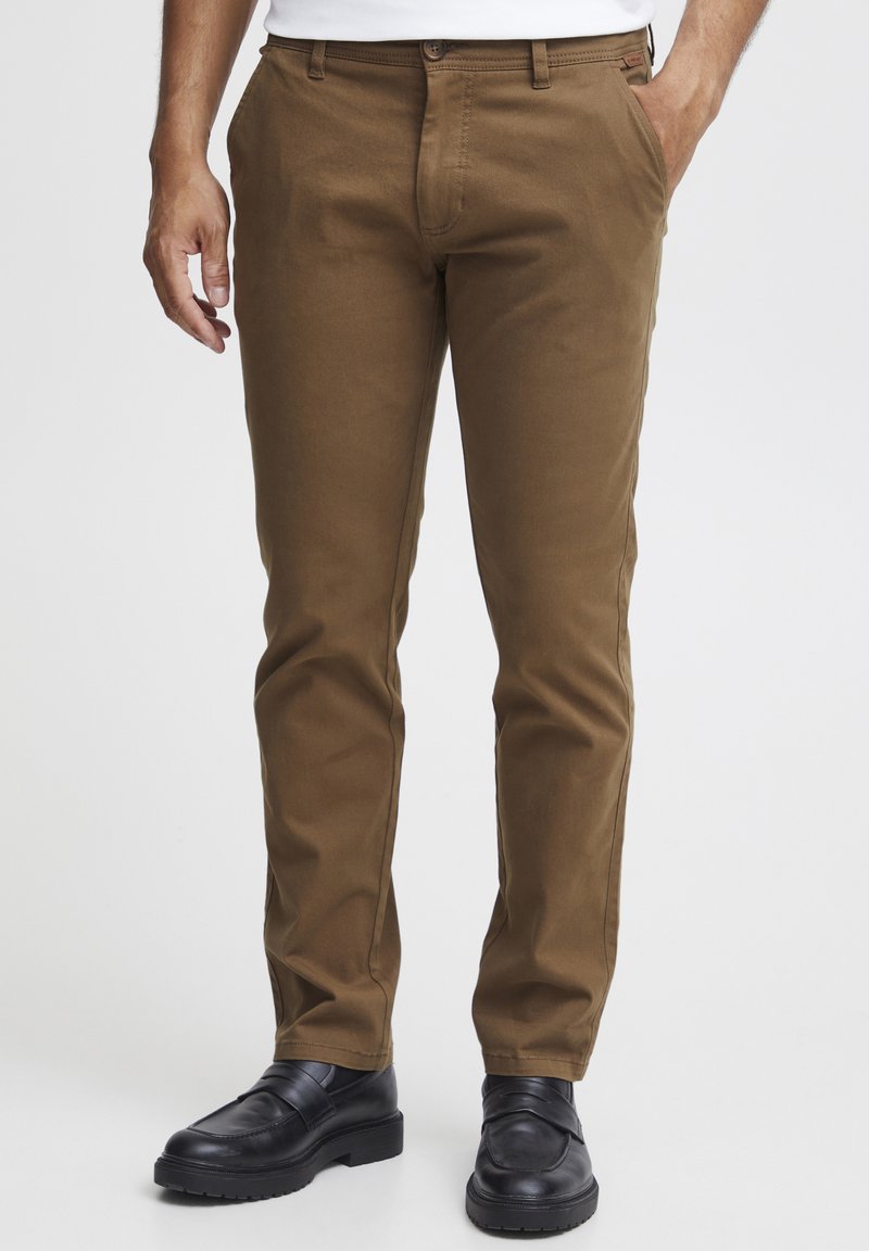 Chinos marrones con una textura de tela suave, corte ajustado y bolsillos frontales. Combinados con zapatos negros sin cordones, con suela plana.