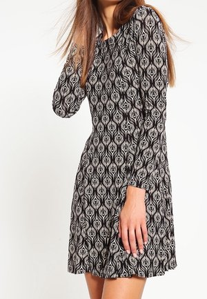 Vrouw die een zwart-witte jurk met lange mouwen draagt met een geometrisch pauwenveermotief, poserend tegen een effen lichte achtergrond.