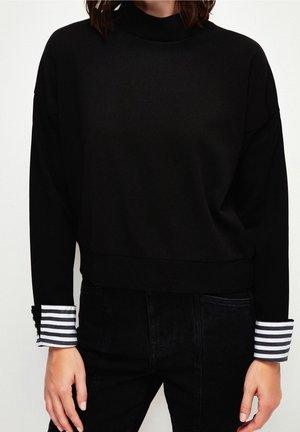 Personne portant un haut noir à manches longues avec des poignets rayés blancs et noirs, associée à un pantalon noir, sur un fond blanc uni.