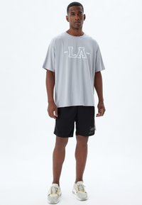 Camiseta atlética gris claro con el logo "LA", de manga corta, combinada con pantalones cortos negros que tienen un parche de logo rectangular, usados con zapatillas claras.