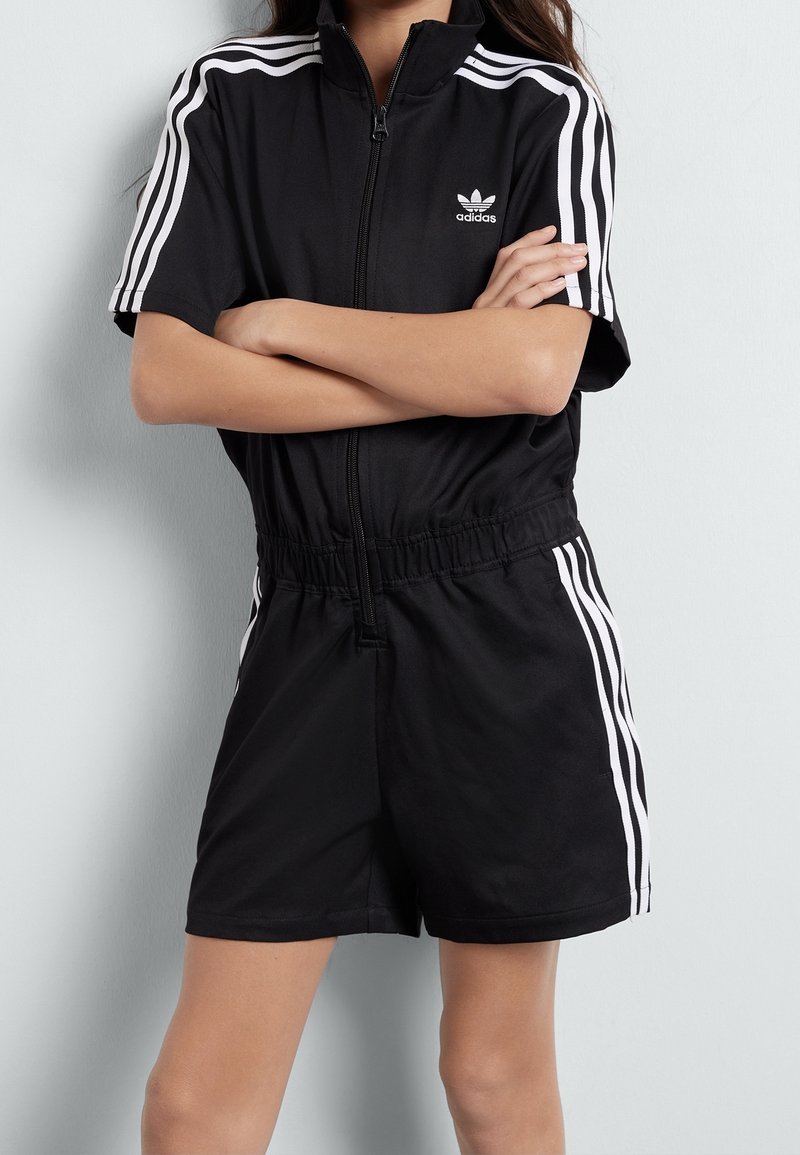 Černý zip-up romper vyrobený z hladké tkaniny, s bílými třemi pruhy na ramenou a elastickým pasem. Přítomné logo Adidas.