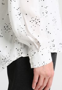 Blusa blanca con patrón de estrellas negras, tela transparente, mangas ligeramente abullonadas y botones decorativos de perlas en el puño.