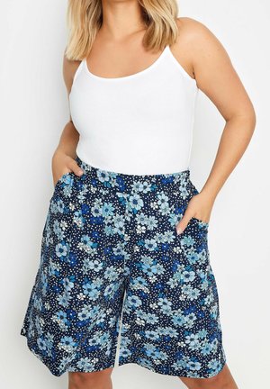 Kvinde iført blå blomstrede knælange shorts med lommer og en hvid ærmeløs top mod en ensfarvet hvid baggrund.