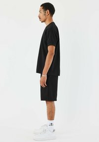 Camiseta negra de manga corta y pantalones cortos a juego, ambos de tejido texturizado. Combinado con zapatillas blancas y calcetines cortos. Vista lateral.