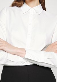 Personne portant une chemise blanche boutonnée avec col et un pantalon noir, les bras croisés sur la poitrine, cadrée sous le menton.