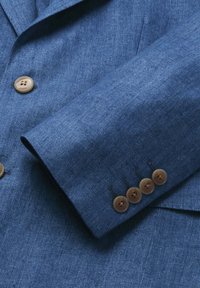 Ärmel eines Blazers aus blau strukturiertem Stoff mit vier braunen Knöpfen und Teil der Vorderseite, die einen passenden Knopf mit Textgravur zeigt.