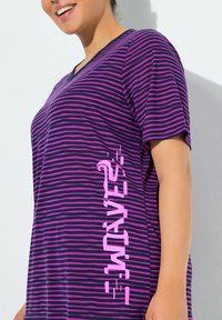 Camiseta de rayas púrpuras y negras con cuello en V y texto rosa intenso en el lateral. Tela suave con mangas cortas y ajuste relajado.
